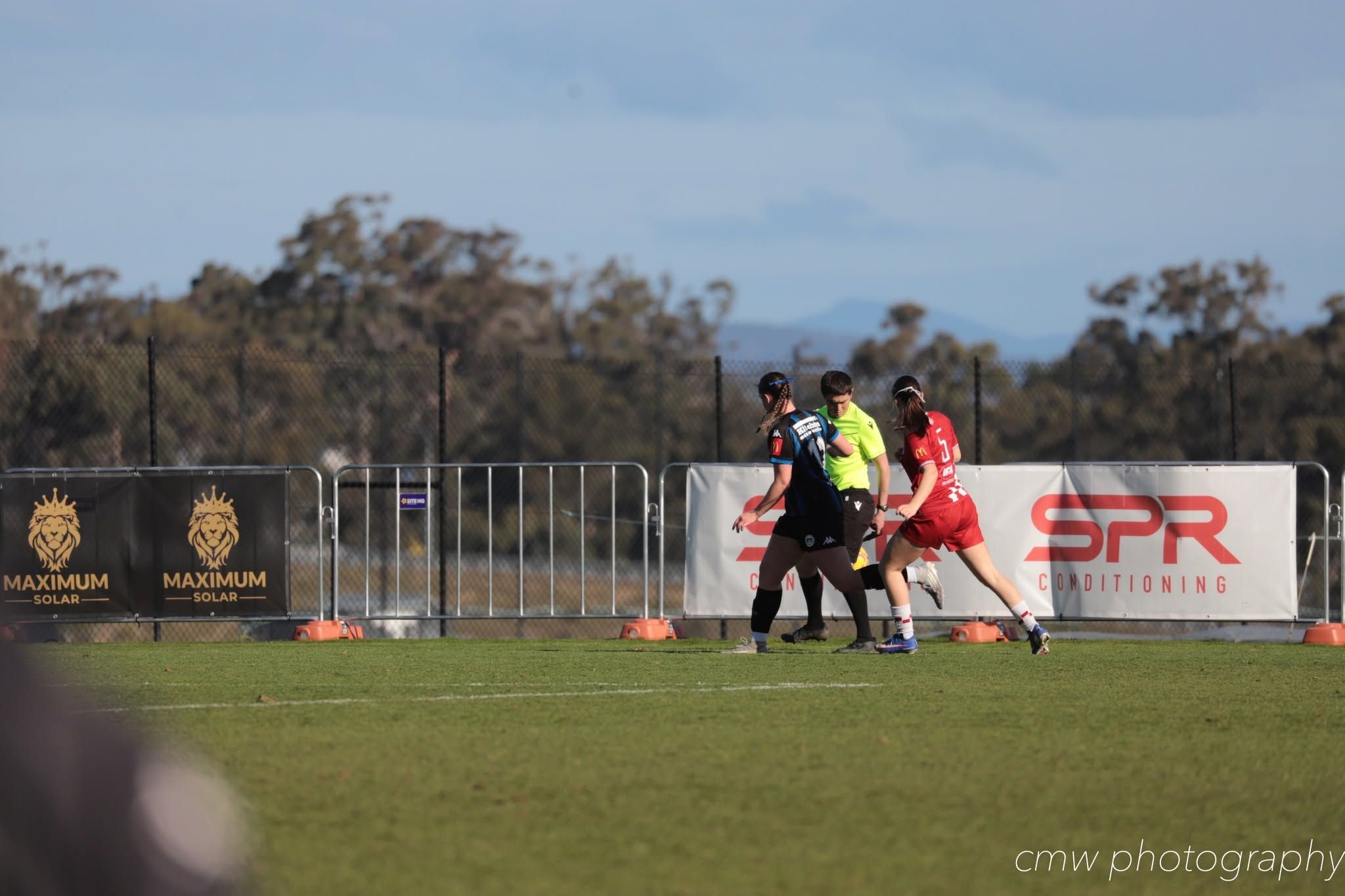 Football players and an umpire beside Maximum Solar banners on the ground fence