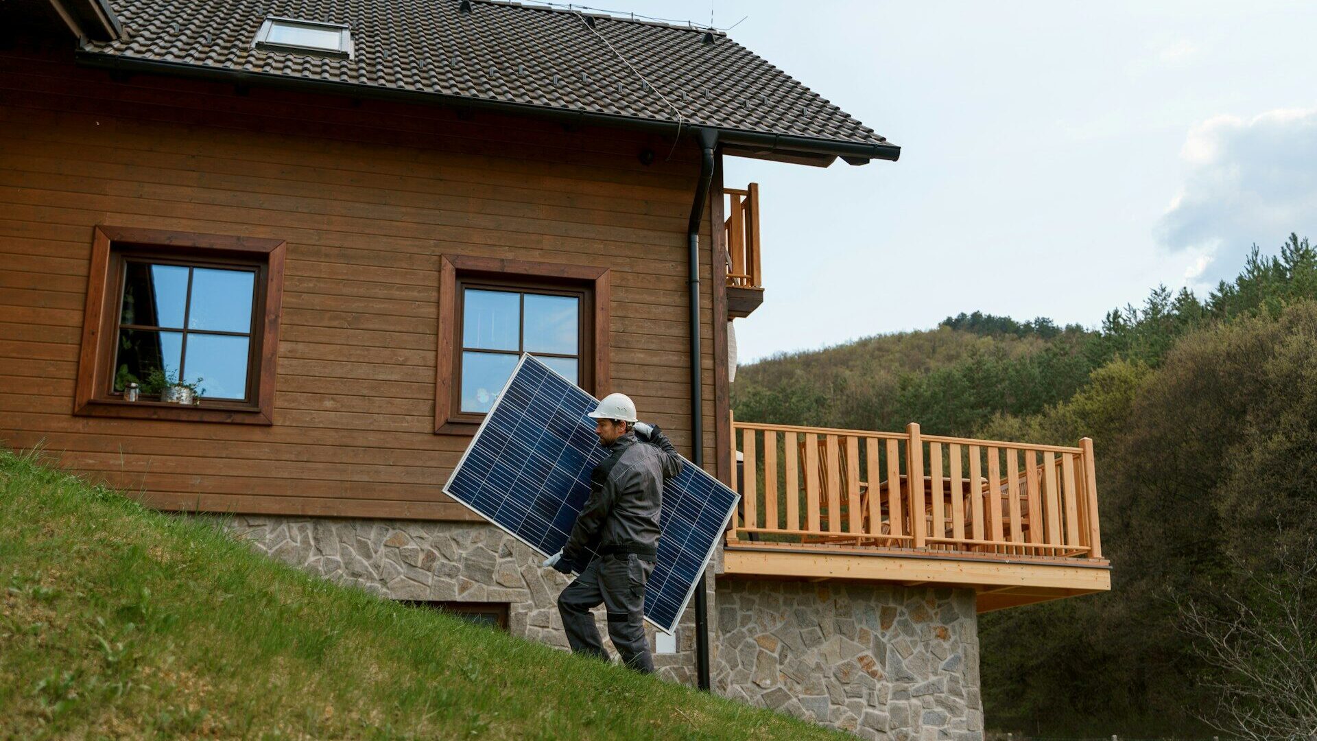 Solar installer carrying panels up a grassy slope beside a rural timber home